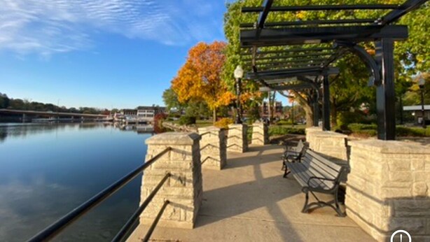 A riverside walkway with a stone railing, a wooden bench, and a pergola with a metal frame.