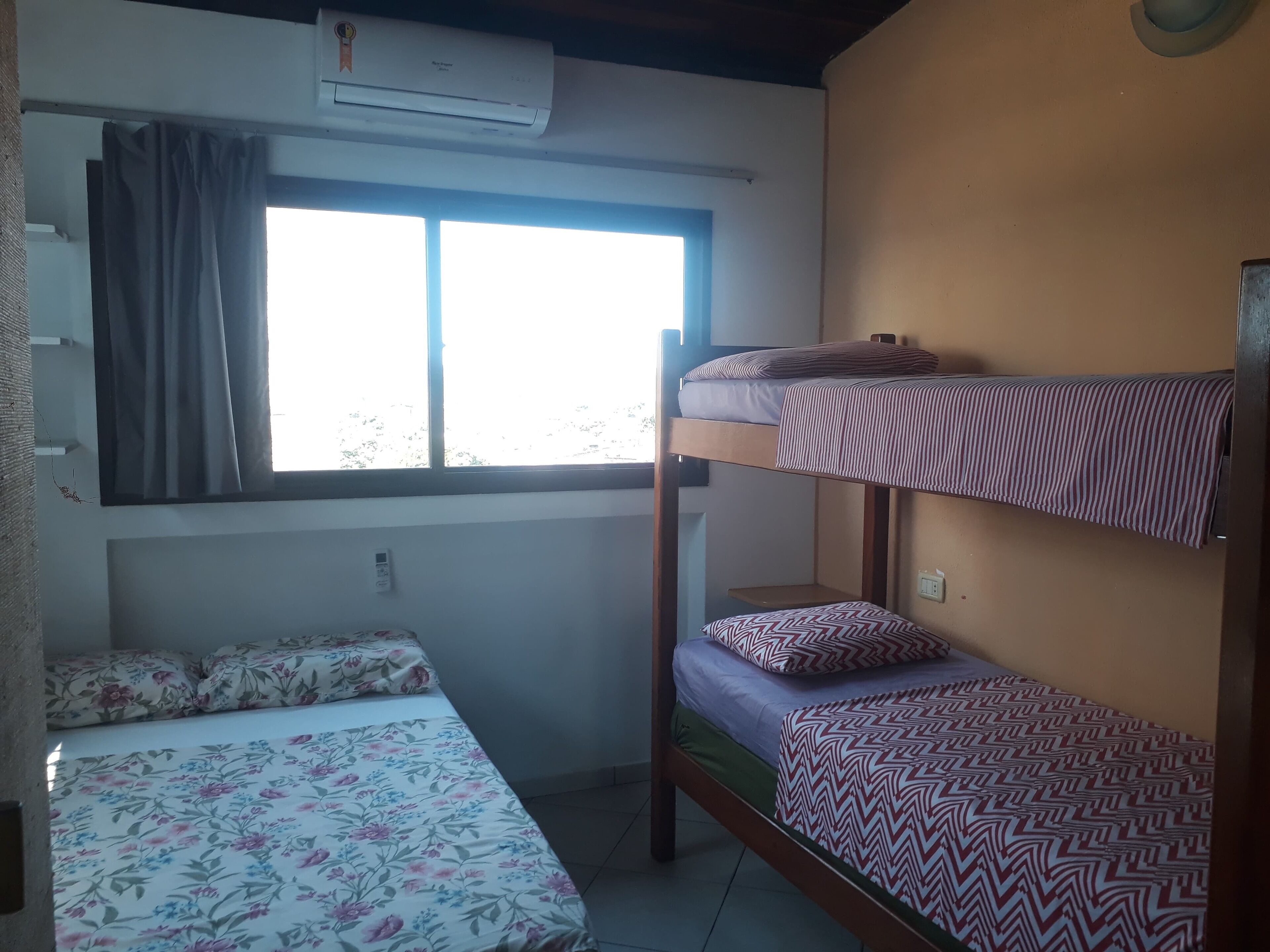 A dormitory room with bunk beds, a window with curtains, an air conditioner, and a thermostat.