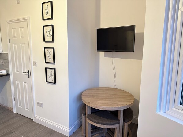 A room with a white door, a round wooden table with two chairs, a mounted flat-screen TV, and four framed artworks on the wall. - Cosy London Apartment by Underground Train station - Elizabeth Line + Parking (Ilford)