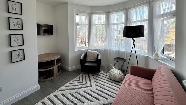 Un salon moderne doté d’une grande fenêtre, d’un canapé noir, d’un canapé rose, d’un téléviseur fixé au mur et d’un meuble de rangement en bois.