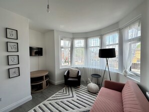 A modern living room with a large window, a black sofa, a pink sofa, a TV mounted on the wall, and a wooden entertainment unit. - Cosy London Apartment by Underground Train station - Elizabeth Line + Parking (Ilford)