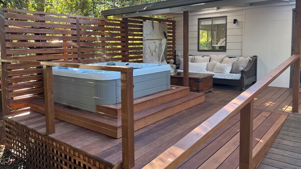 Une terrasse en bois avec un jacuzzi, un canapé et un escalier menant à un espace abrité doté d’une fenêtre et d’un miroir.