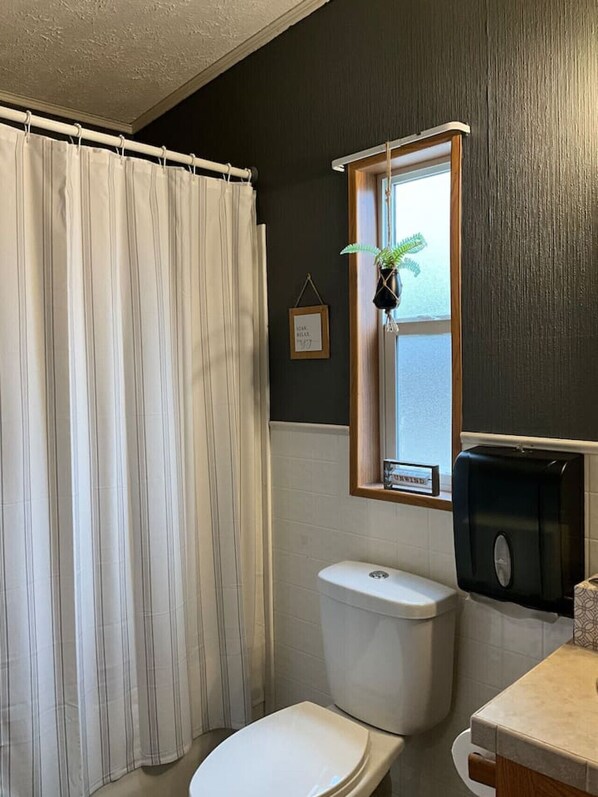 A bathroom with a white toilet, a black soap dispenser, a window with a plant, and a shower curtain.