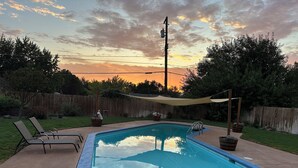 A rectangular swimming pool with a blue edge, surrounded by a concrete deck. There are two lounge chairs and a small table with a statue. A wooden fence and trees are in the background. - Clarkston House with pool! Relax and enjoy with family and friends (Clarkston)