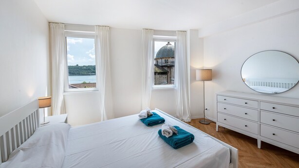 Hotelkamer met een bed, twee ramen, een dressoir, een lamp en uitzicht op een gebouw en water.