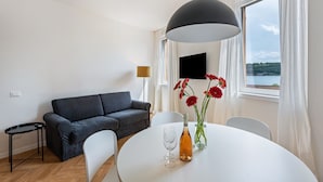 Een moderne kamer met een grijze bank, een ronde eettafel met een fles en glazen, een televisie aan de muur en een raam met uitzicht.