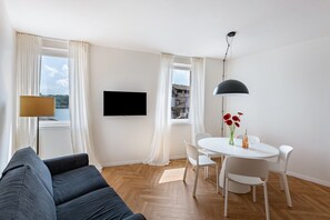 Una sala de estar moderna con un sofá, una mesa de comedor con sillas, un televisor montado en la pared y vistas a un edificio a través de las ventanas.
