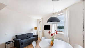 Een moderne woonkamer met een bank, een eettafel met stoelen, een aan de muur bevestigde televisie en een raam met gordijnen.