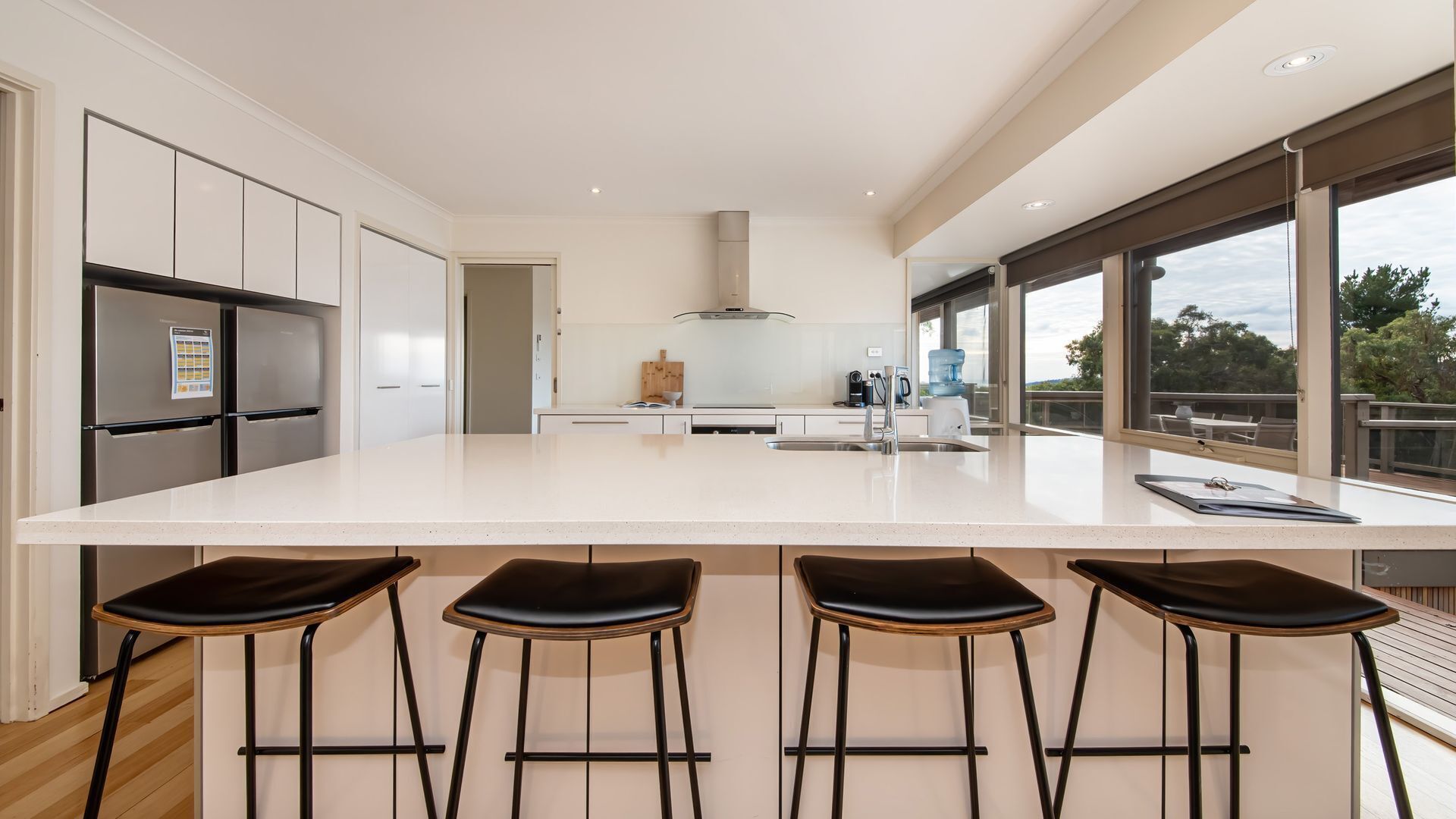 Une cuisine moderne dotée d’un grand îlot central, de tabourets de bar, d’appareils électroménagers en acier inoxydable et de grandes fenêtres offrant une vue sur l’extérieur.