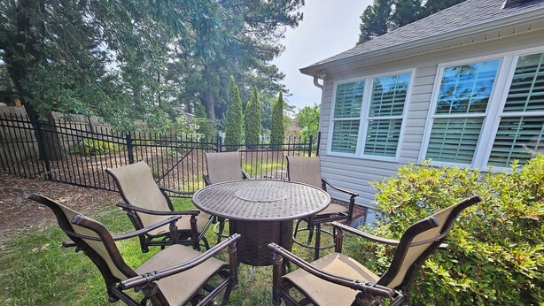 Een terras met een ronde tafel en vier stoelen, omgeven door groen en een huis op de achtergrond.