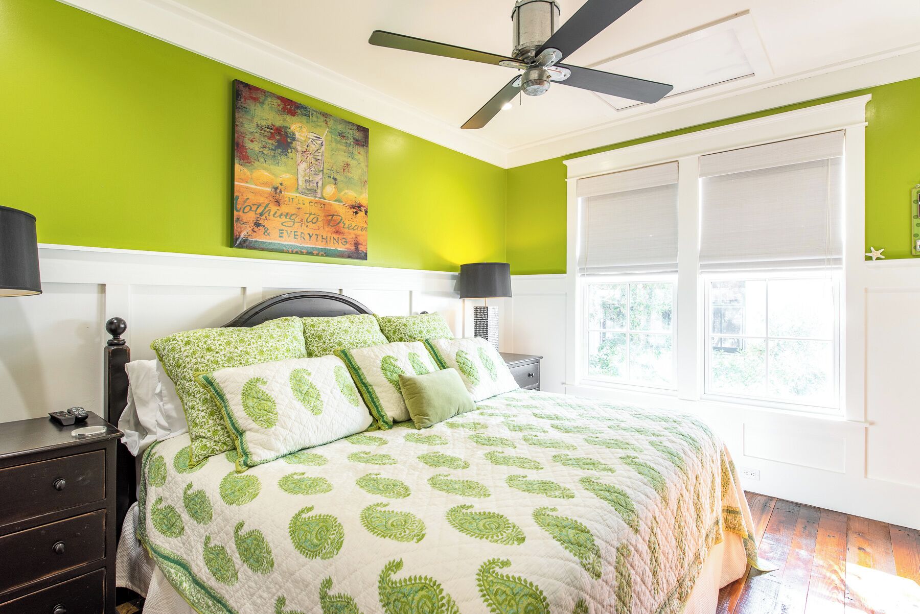 A bedroom with a bed, a nightstand, a lamp, a ceiling fan, a window, and a painting on the wall.