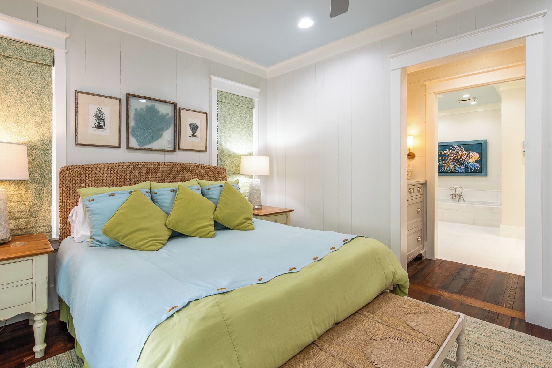 Un dormitorio con cama, mesitas de noche, lámpara, cuadro en la pared y un baño visible a través de una puerta abierta.