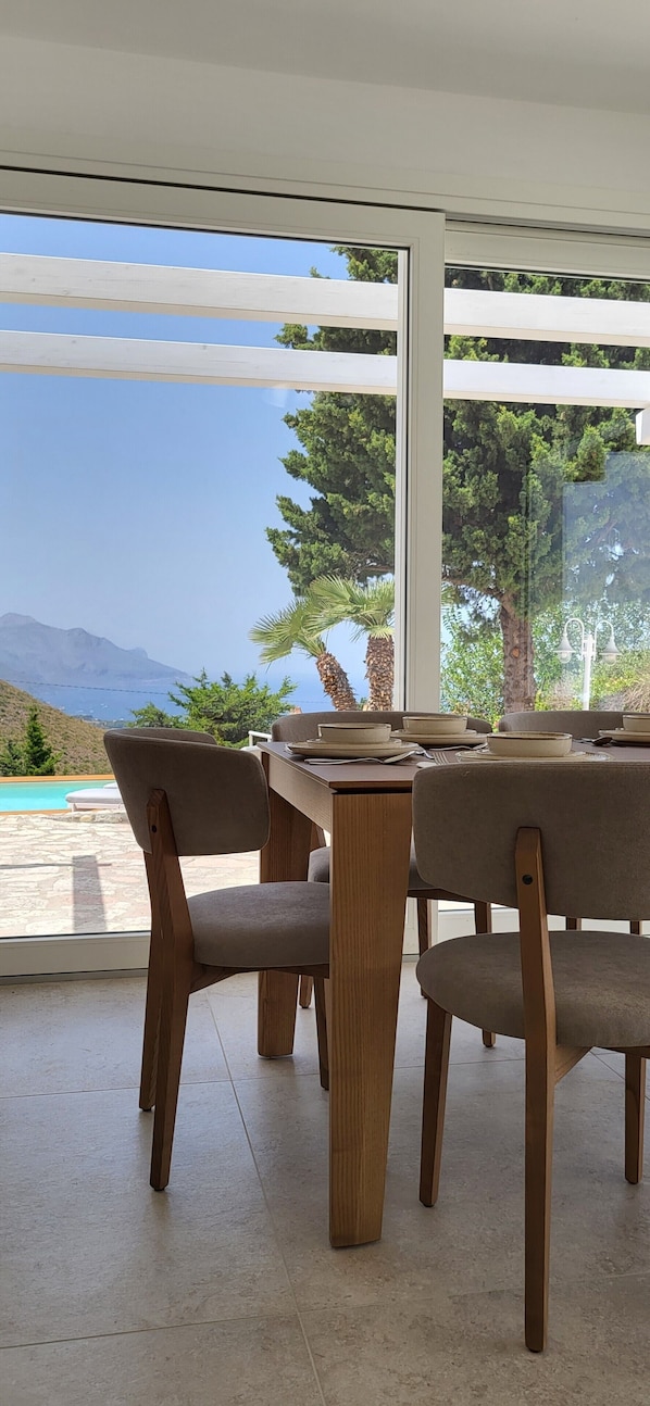A dining area with a table and chairs, a view of the sea and mountains, and a pool outside. - Villa 'Ballerina Bianca' with Private Pool, Wi-Fi and Air Conditioning (Scopello)