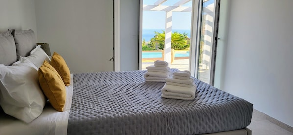 Un dormitorio con cama, almohadas, y ventana con vista a una piscina y follaje.