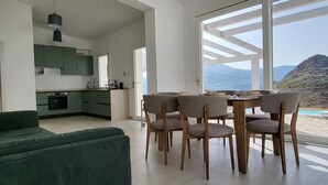 Un espacio habitable moderno con sofá verde, mesa de comedor con sillas, cocina y un ventanal con vistas a la montaña.