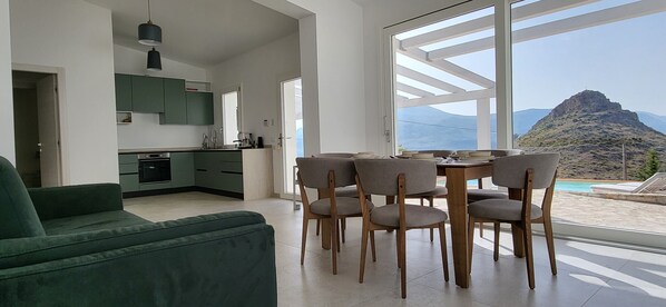 A modern living space with a green sofa, dining table with chairs, a kitchen, and a large window with a mountain view. - Villa 'Ballerina Bianca' with Private Pool, Wi-Fi and Air Conditioning (Scopello)