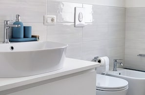 Een badkamer met een witte wastafel, toilet en badkuip. Op de wastafel staat een handdoekrek en een zeepdispenser.