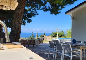 Un espace extérieur aménagé avec des sièges, offrant une vue sur la mer, une table avec des chaises et un grand arbre.