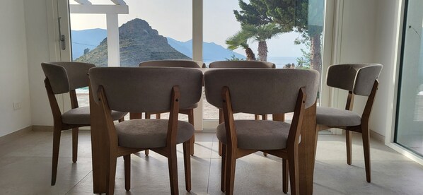 Een kamer met uitzicht op bergen en de zee, met vier houten stoelen en grijze kussens.