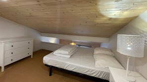Ein Schlafzimmer mit schrägen Wänden, einem Bett mit weißer Bettwäsche, einem Nachttisch mit Lampe und einer Kommode.