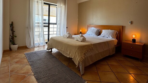 A bedroom with a bed, a nightstand, a lamp, a window with curtains, and a potted plant. - Apartamento Mar Olhão | Spacious 2-Bedroom Apartment in Prime Location (Olhão)