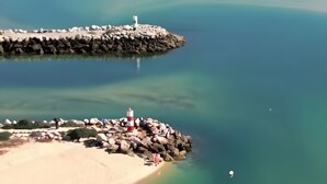 A coastal landscape with clear waters, a sandy beach, and a rocky breakwater with a lighthouse. - Apartamento Mar Olhão | Spacious 2-Bedroom Apartment in Prime Location (Olhão)