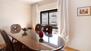 A dining area with a glass table, chairs, a bowl, and a window with curtains. - Apartamento Mar Olhão | Spacious 2-Bedroom Apartment in Prime Location (Olhão)