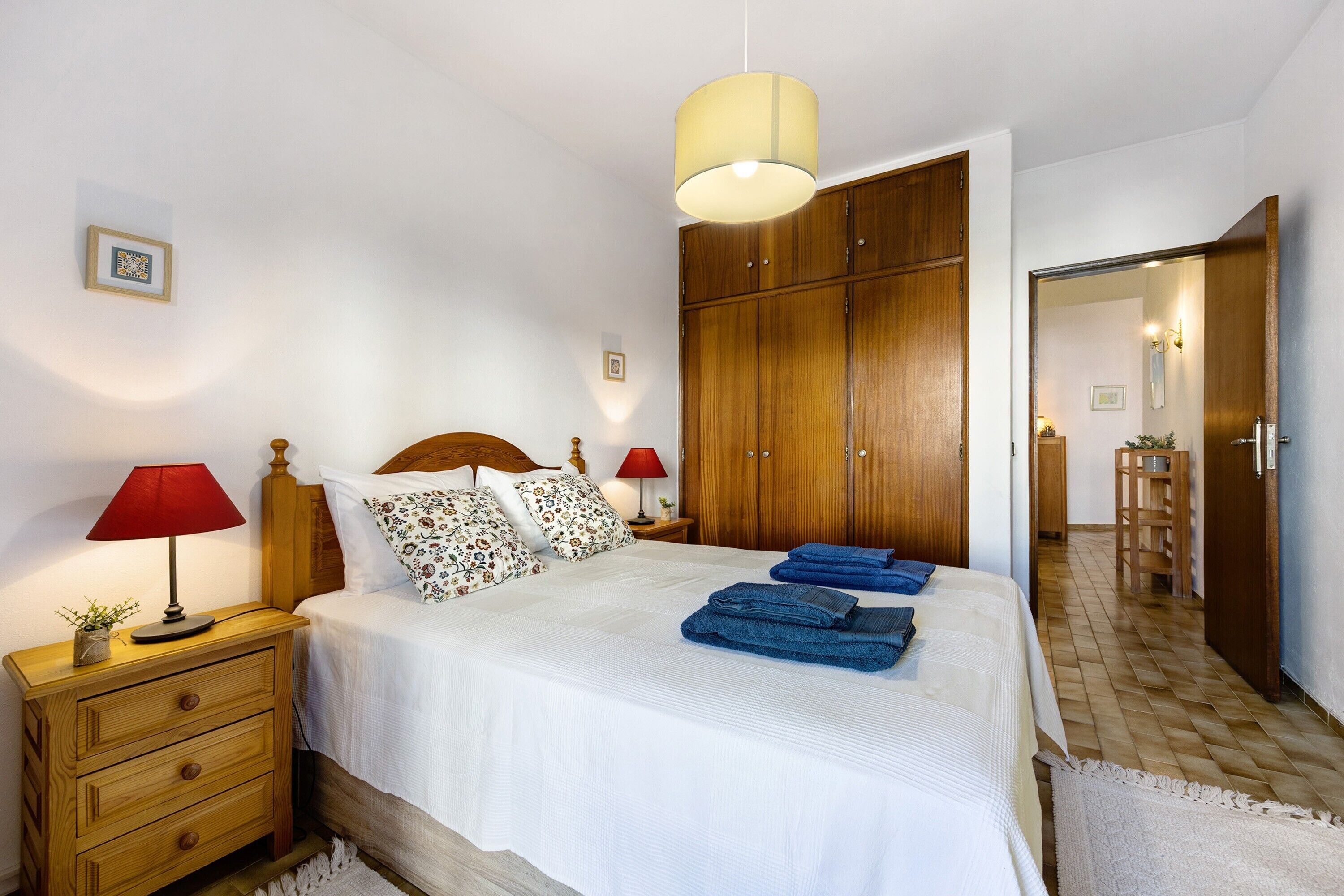 A bedroom with a bed, nightstands, a wooden wardrobe, and a hallway leading to a bathroom.