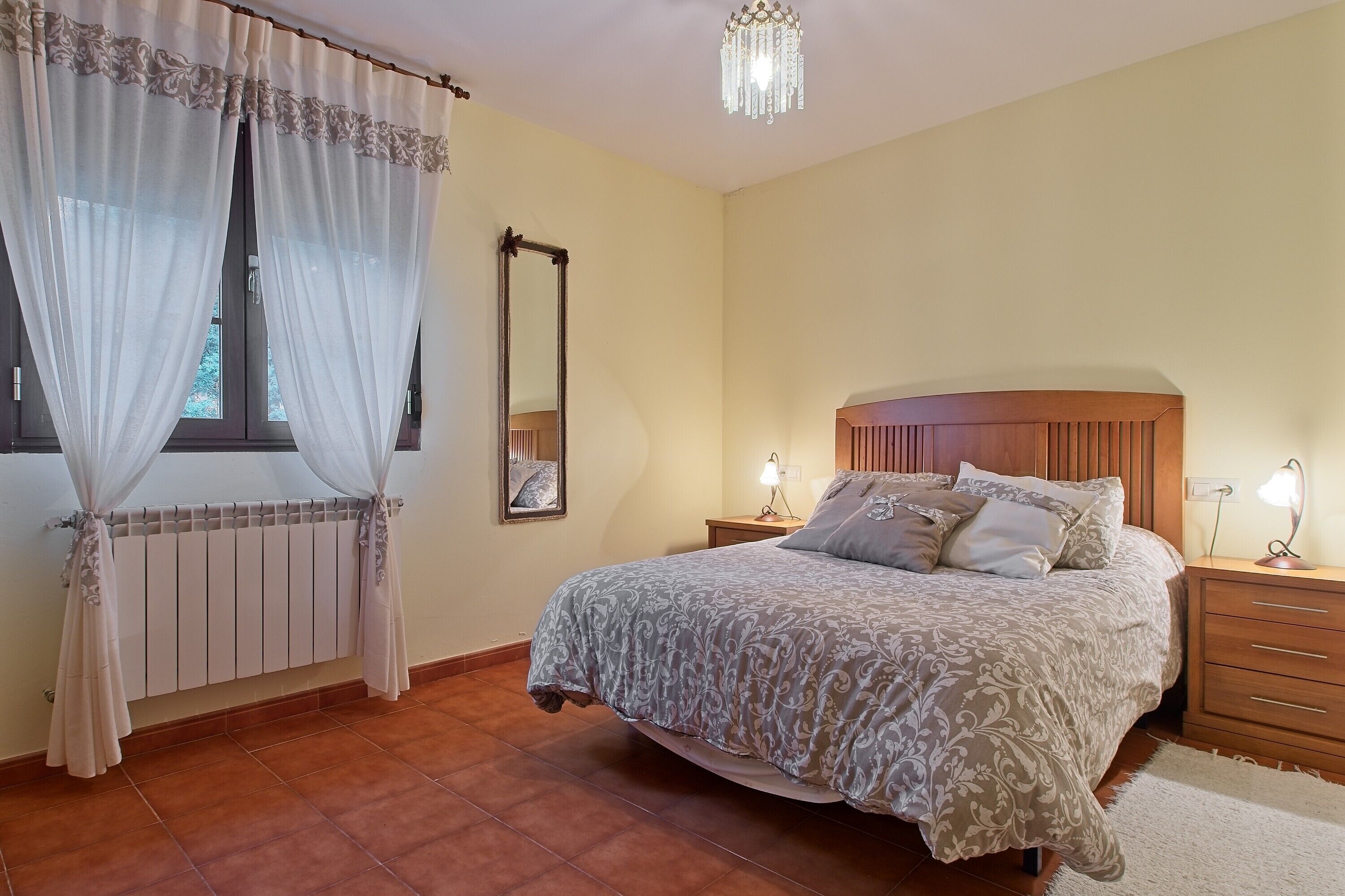 Ein Schlafzimmer mit Bett, Nachttischen, einem Spiegel, einer Pendelleuchte und einem Fenster mit Vorhängen.
