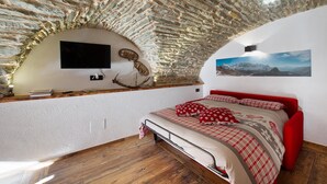A bedroom with a stone ceiling, a mounted TV, a bed with red bedding, and a mountain landscape painting. - Apartment 'La Cachette' with Mountain View, Private Terrace and Wi-Fi (Aosta)