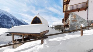 A snow-covered building with a stone wall and wooden balcony, surrounded by mountains. - Apartment 'La Cachette' with Mountain View, Private Terrace and Wi-Fi (Aosta)
