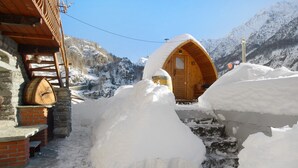A wooden cabin with a snow-covered entrance, a stone wall, and a wooden barrel. - Apartment 'La Cachette' with Mountain View, Private Terrace and Wi-Fi (Aosta)