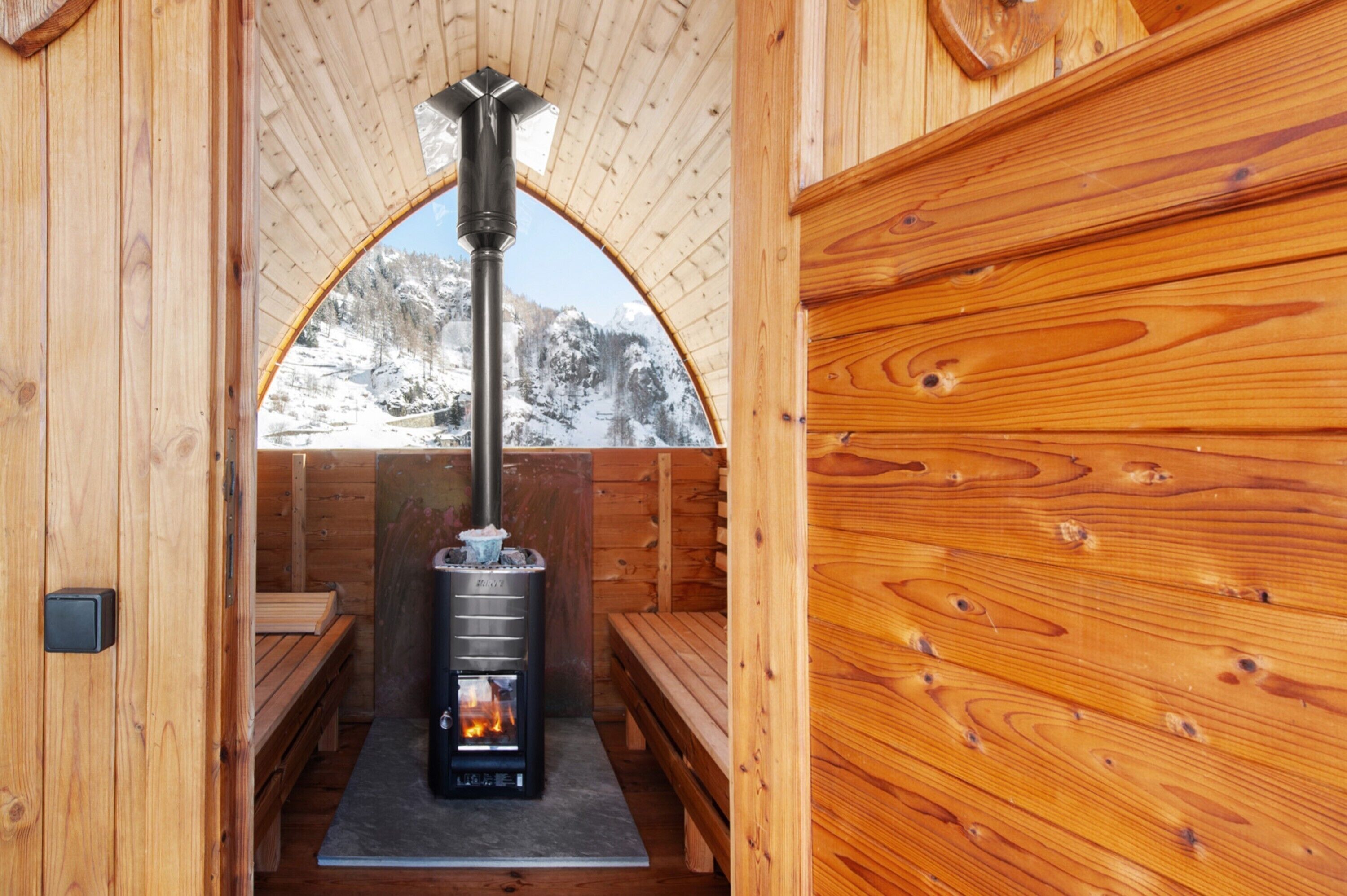 A wooden sauna with benches, a central heating unit, and a view of snow-covered mountains.
