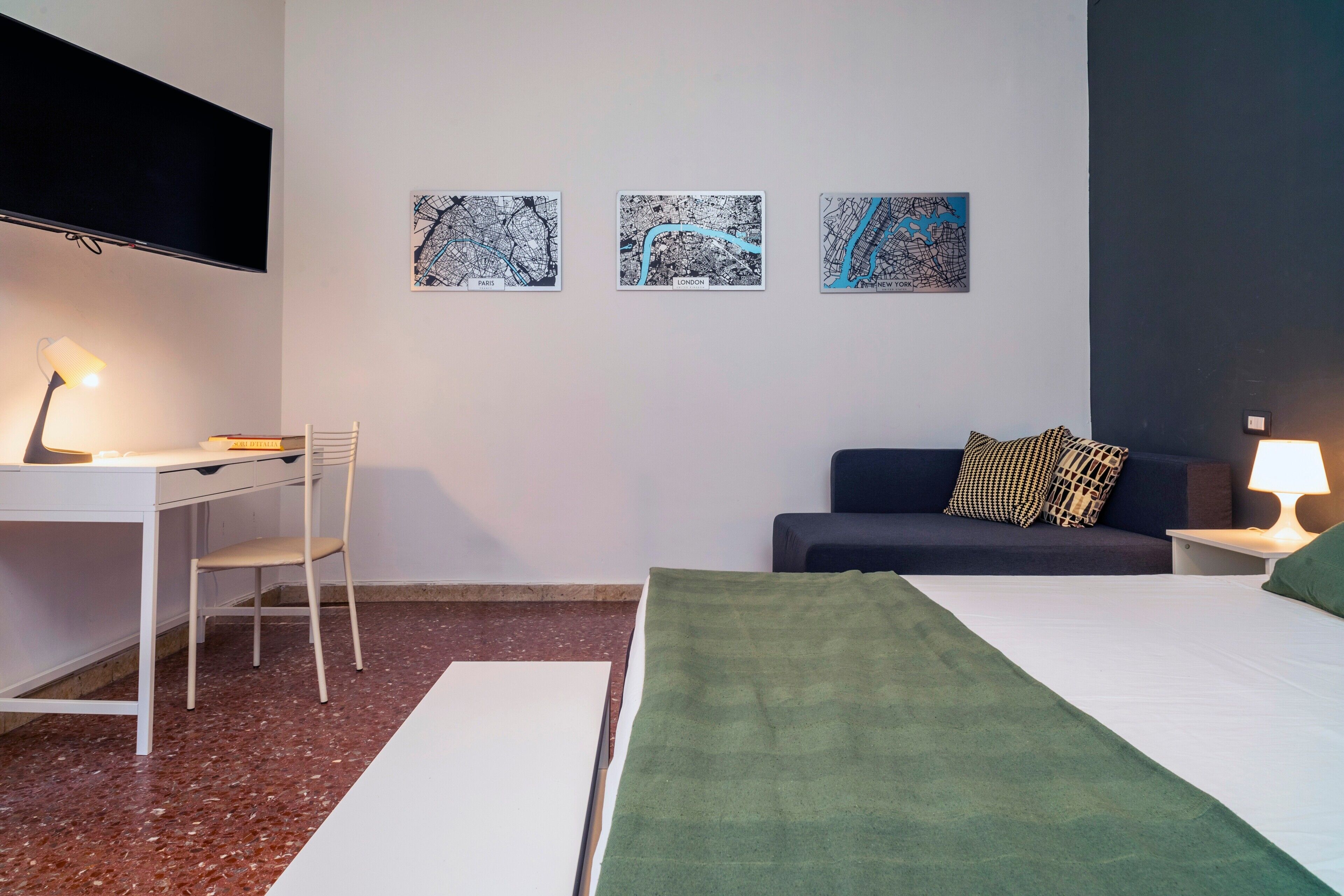 Hotellihuone, jossa on sänky, työpöytä, tuoli, seinälle asennettu televisio ja kolme kehystettyä karttaa seinällä.