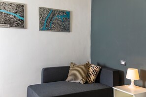 Una stanza con un divano grigio, due cuscini decorati, una lampada a parete e due mappe incorniciate di Londra e New York.