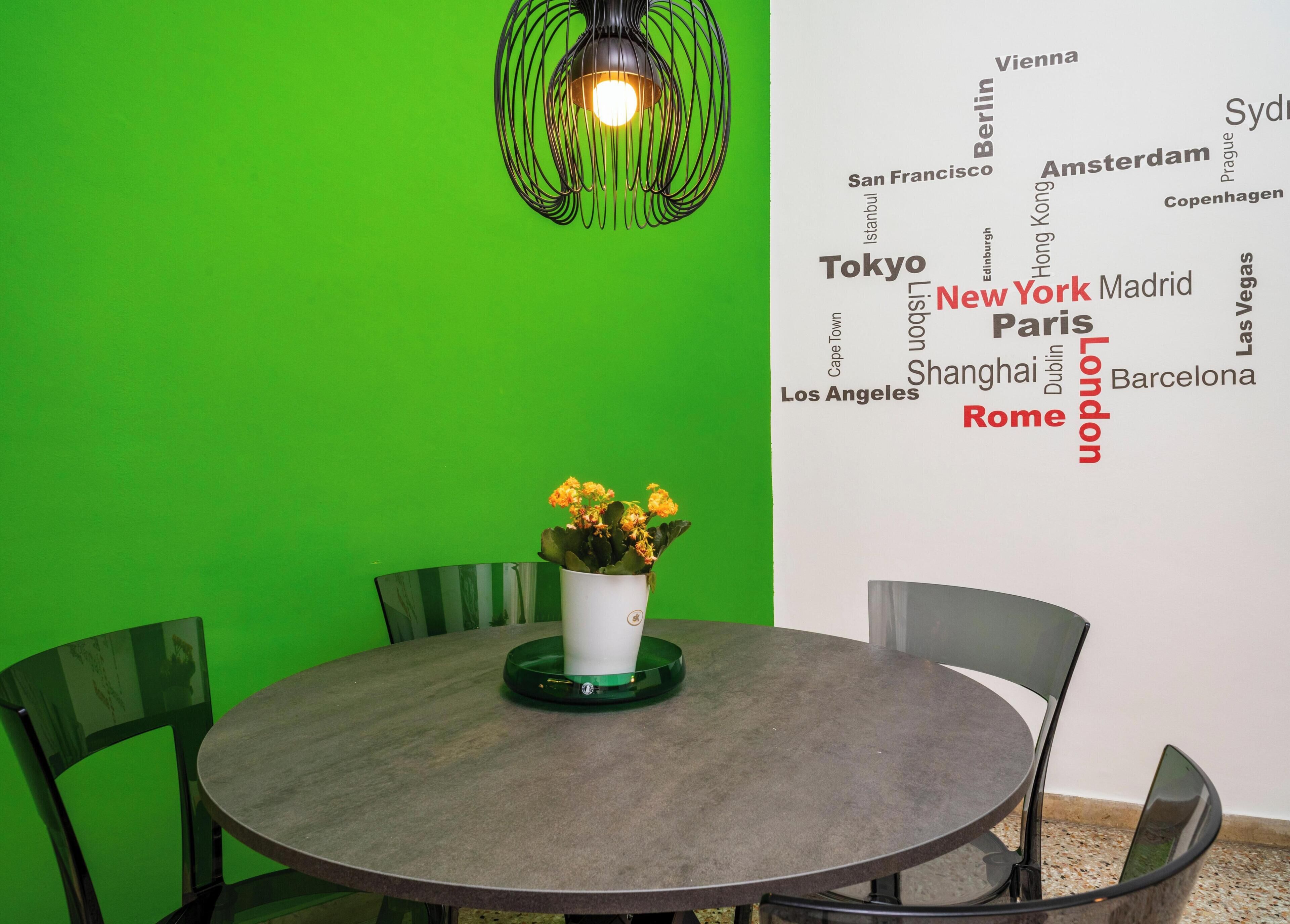 A round table with chairs, a potted plant, and a wall with city names.