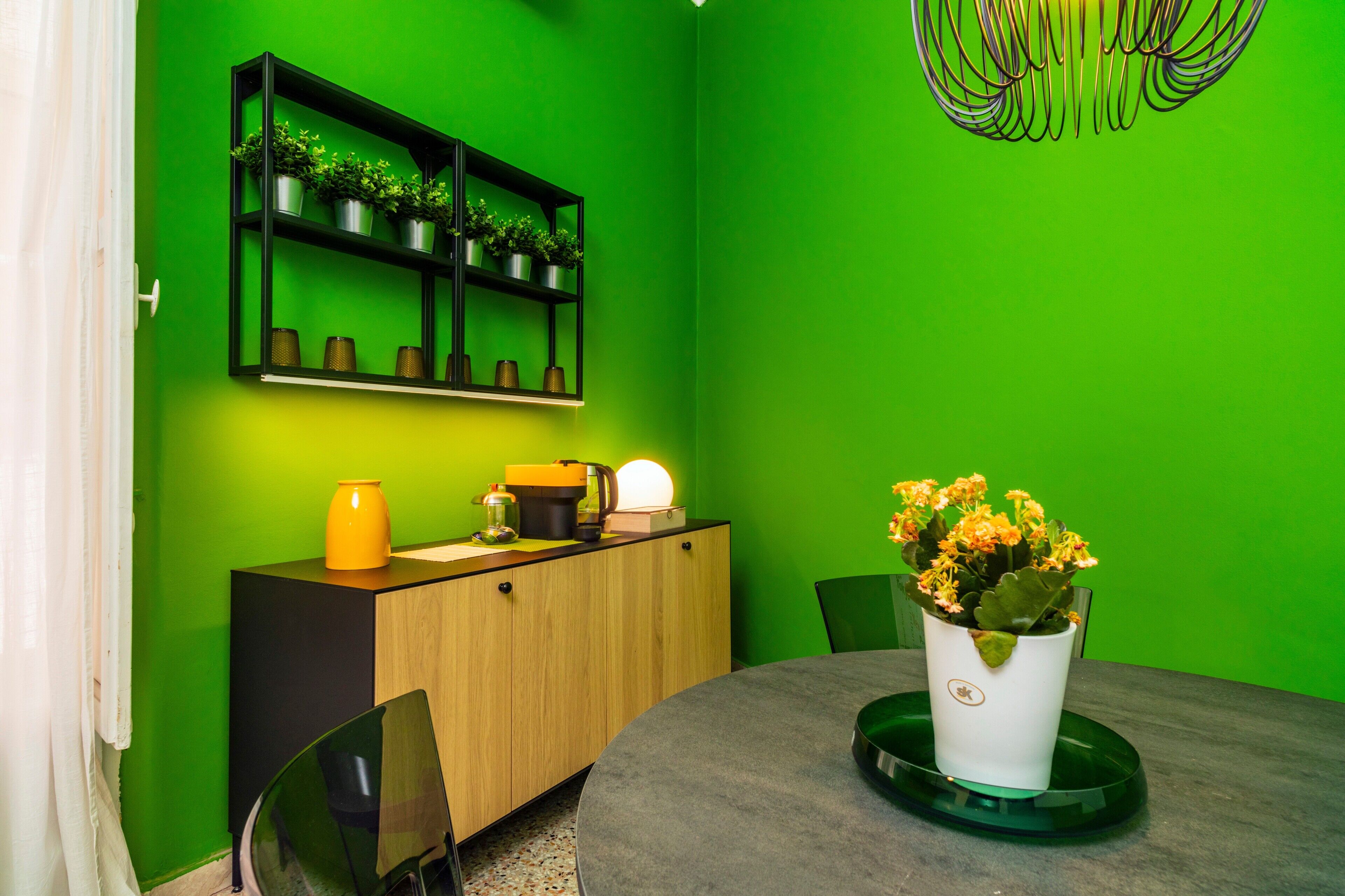 Una habitación con pared verde, un armario de madera, una mesa redonda con planta en maceta y una estantería negra con plantas.