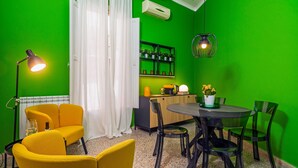 A room with green walls, a dining table with chairs, a yellow sofa, a floor lamp, and a kitchen area with shelves.