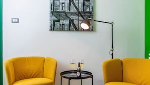 A room with two yellow chairs, a small round table with two glasses and a bottle, a floor lamp, and a wall-mounted artwork.