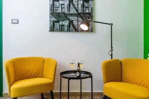 Une pièce avec deux chaises jaunes, une petite table ronde avec deux verres et une bouteille, un lampadaire et une œuvre d’art accrochée au mur.