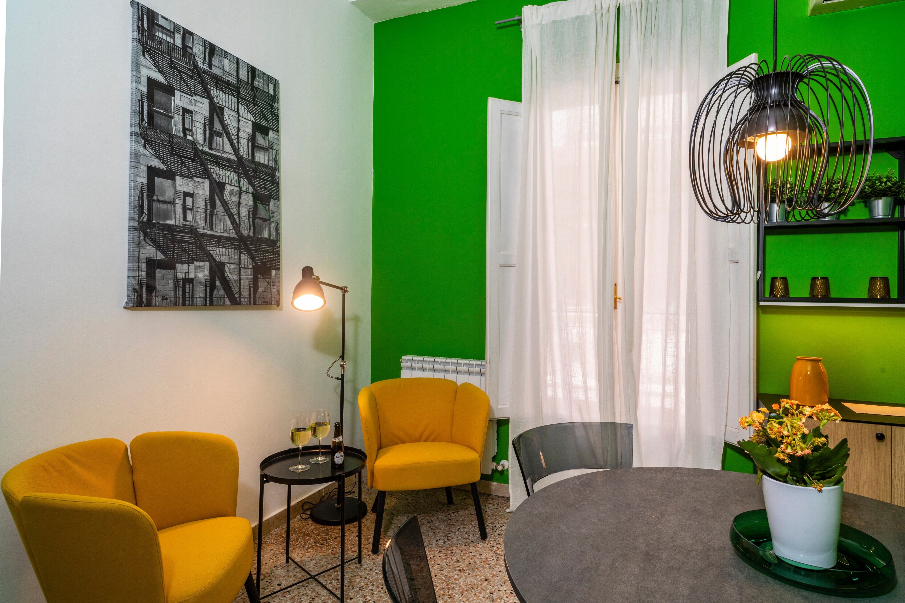 Une pièce avec un mur vert, un tableau en noir et blanc, un fauteuil jaune, une table ronde, un lampadaire et une plante en pot.