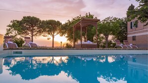 Une piscine bordée de chaises longues et agrémentée d’un belvédère, le tout entouré d’arbres et baigné par les couleurs d’un coucher de soleil.