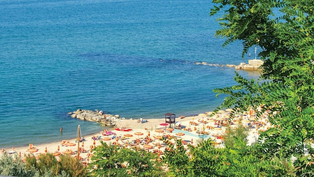 Une plage dotée de nombreux parasols et de chaises longues, un rivage rocheux et une vue dégagée sur la mer.