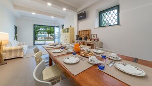 Un coin repas avec une longue table en bois dressée pour le petit-déjeuner, un canapé et un espace cuisine équipé d’une télévision et d’une fenêtre.