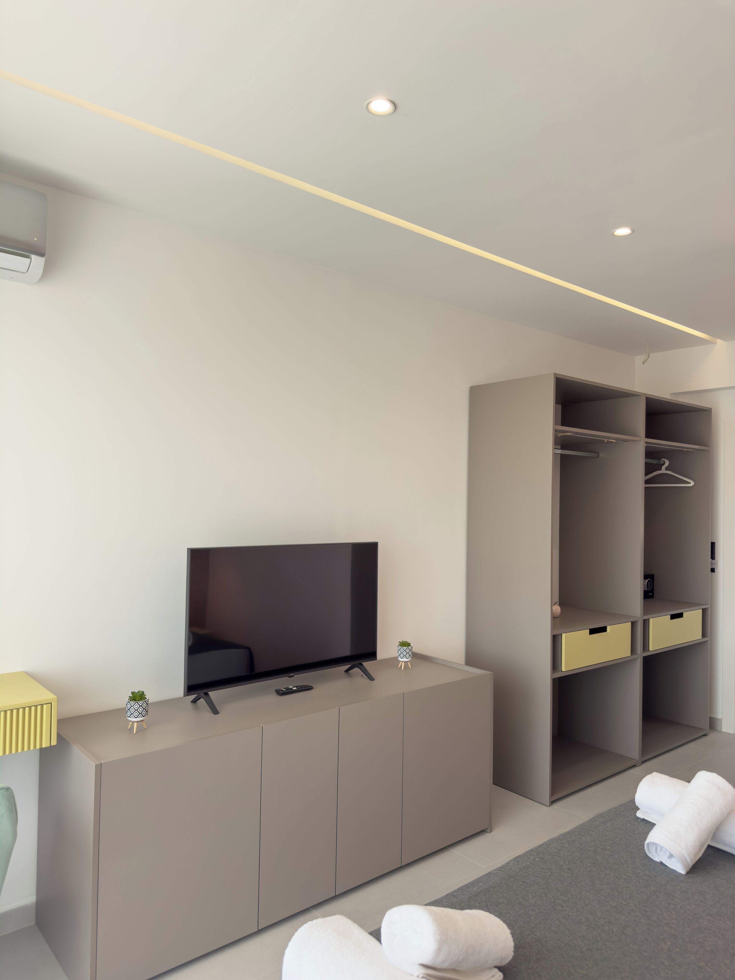 A modern hotel room with a flat-screen TV on a cabinet, a walk-in closet with shelves, and a grey carpet.