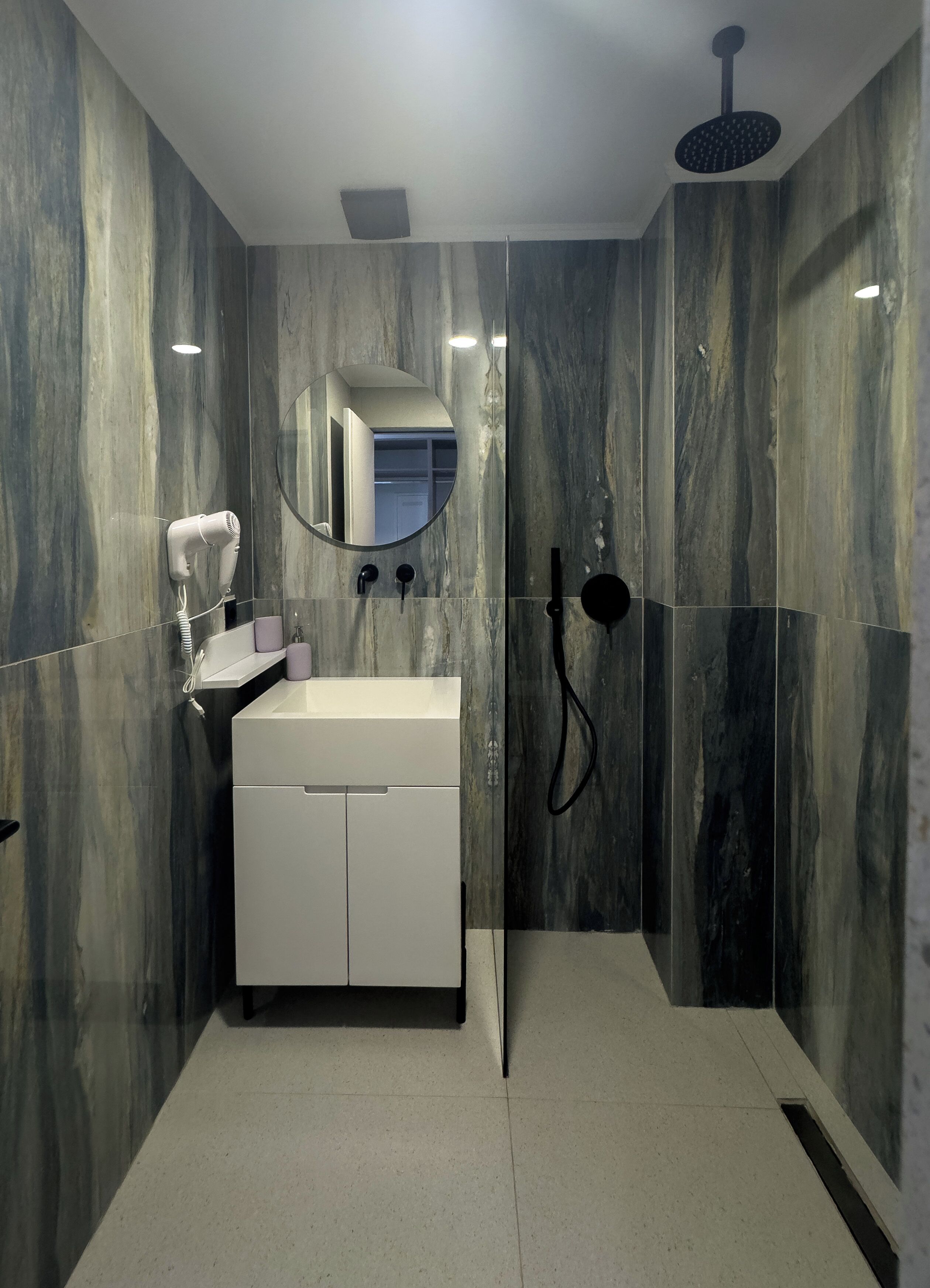 Un baño moderno con tocador blanco, espejo, secador de pelo y ducha con manguera negra.