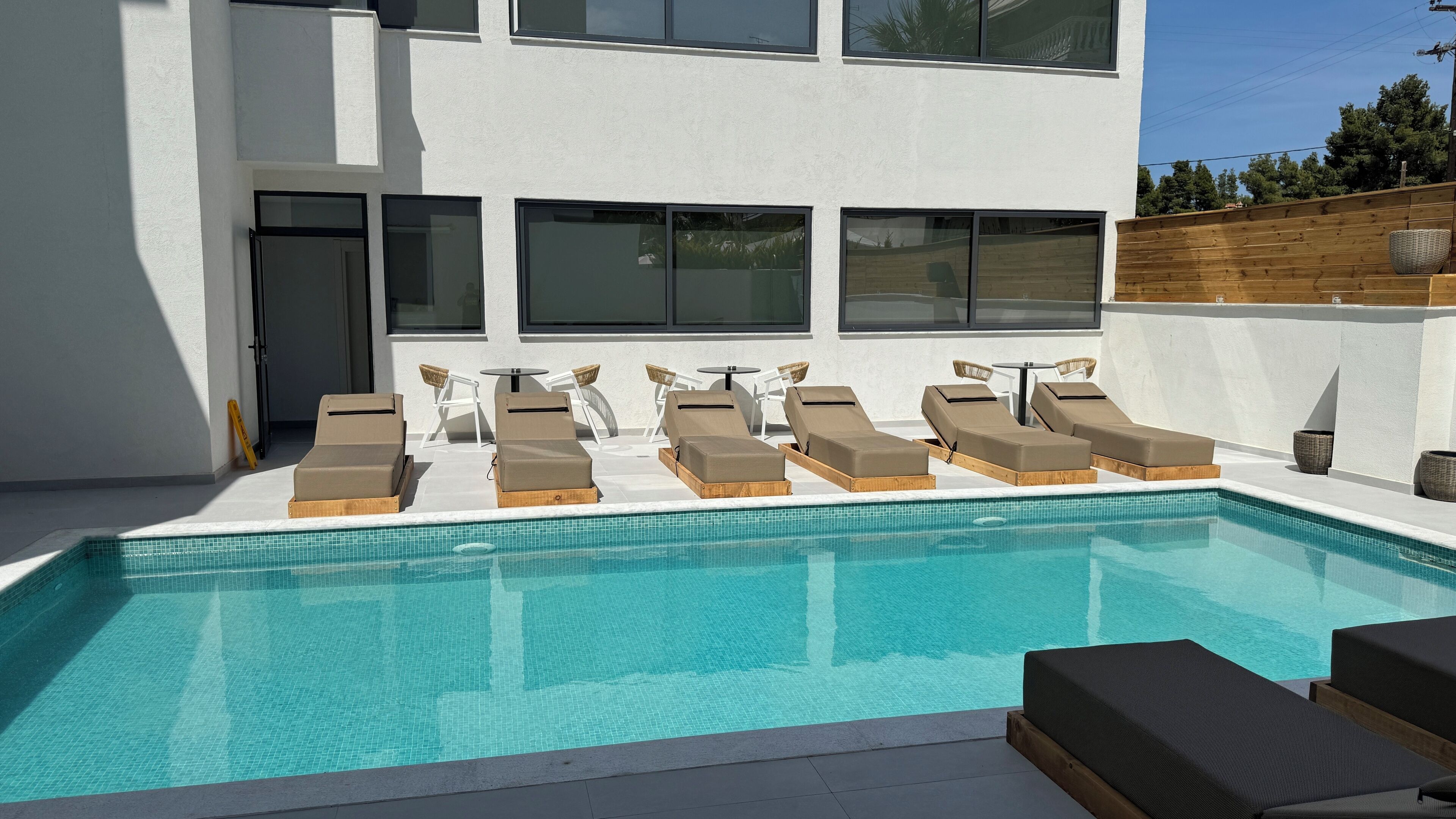 A modern pool area with multiple sun loungers and a small table, surrounded by large windows and a wooden fence.