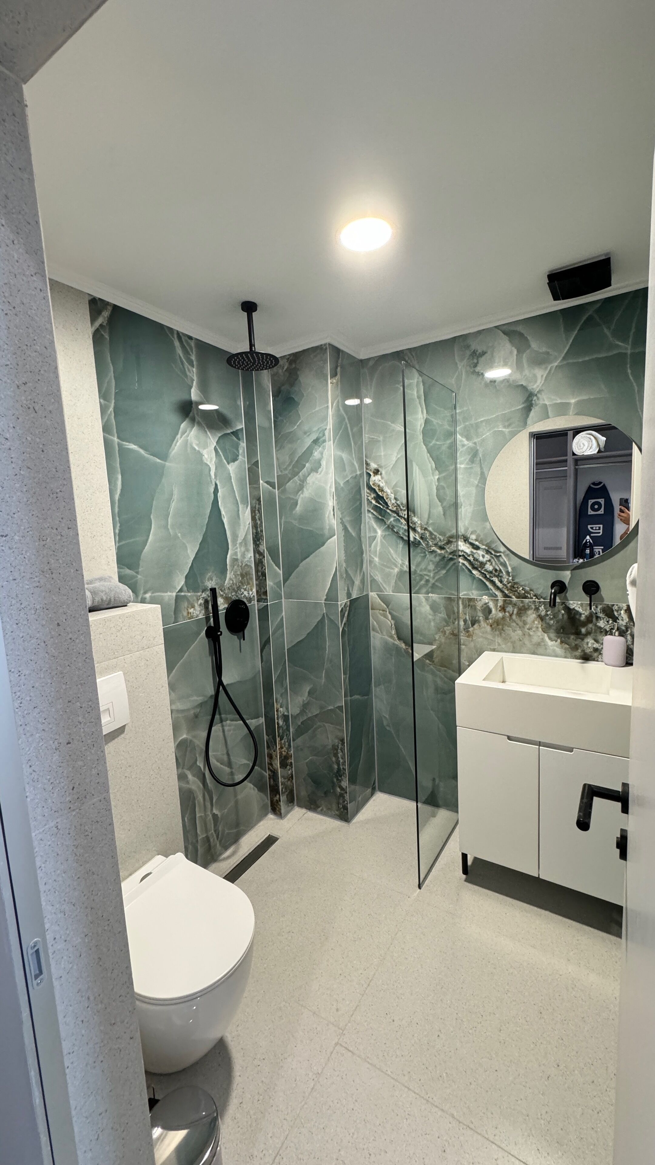 A modern bathroom with a marble wall, a glass shower enclosure, a white sink, and a toilet.