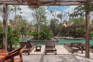 Un área junto a la piscina con sillones y mesas de madera, rodeada de exuberante vegetación y árboles.