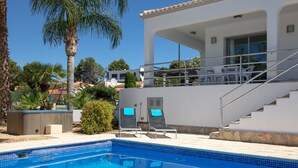 Una piscina con borde de azulejos, un área de estar techada al aire libre con sillones de descanso, y una escalera que conduce a un balcón en un segundo piso.